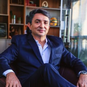 Foto do autor. Homen de cor morena de terno sentado em uma poltrona de blaser azul escuro aberto e camisa azul clara